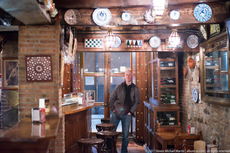 Man standing in a rustic Granada tavern with timber beams, brick walls, wooden bar, stools, and ceramics.