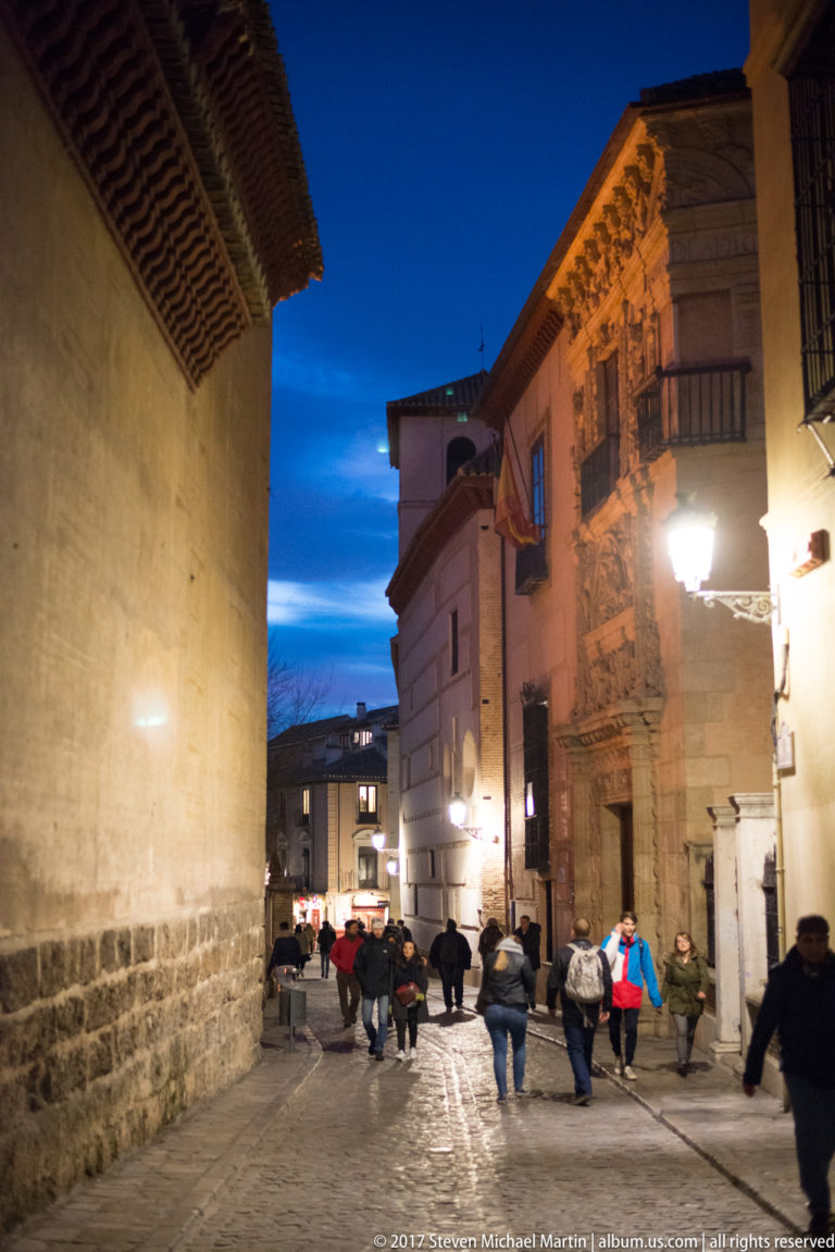 Pedestrians on a narrow Granada street beside an illuminated, richly carved Renaissance stone façade at dusk.