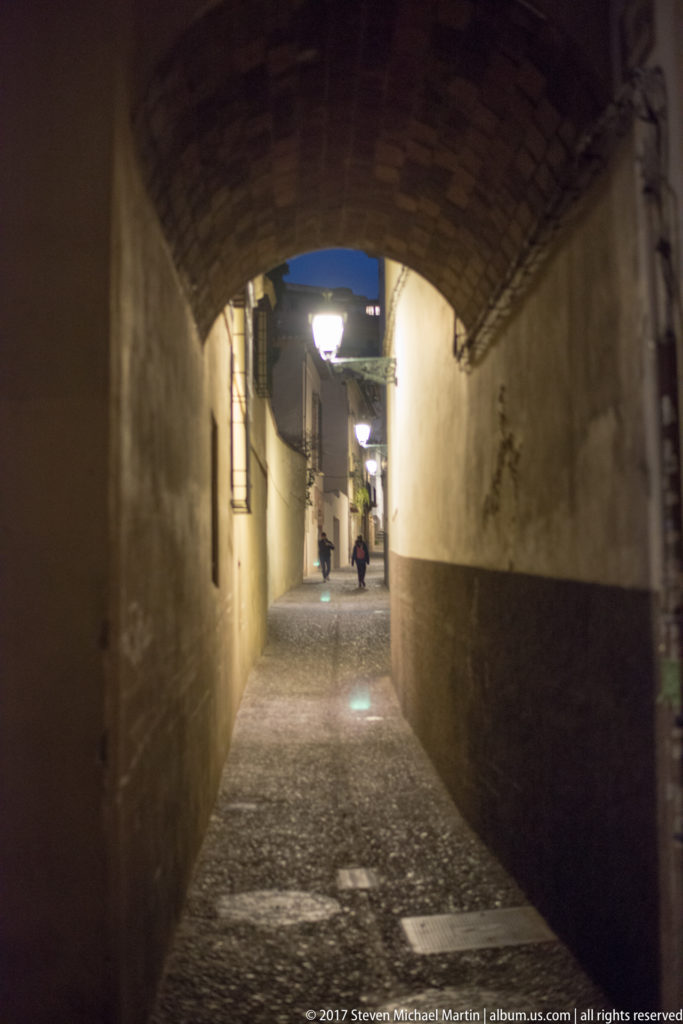 Narrow cobbled passage beneath a low masonry arch, lit by wall lamps with pedestrians in the distance.