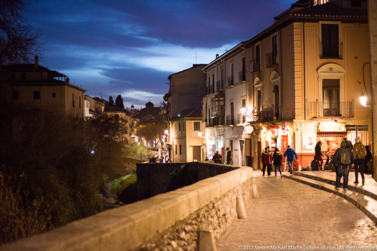 Pedestrians on the stone-paved Carrera del Darro beside a parapet and historic buildings at dusk.