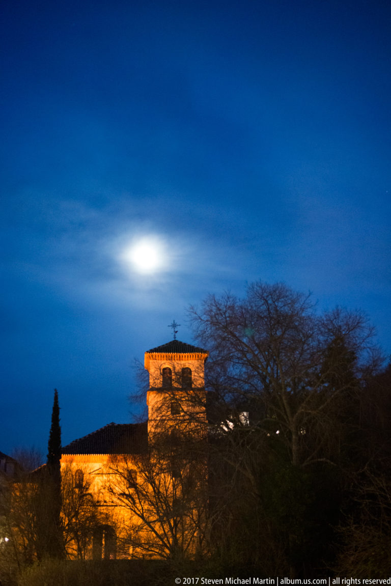 Amber-lit historic building with tiled roof and square upper section beneath a bright moon in Granada.