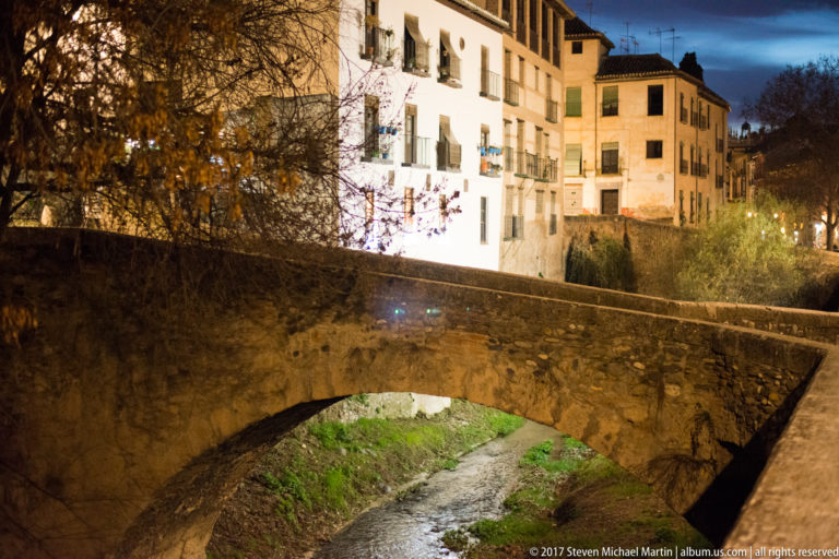 Single-arch stone bridge over the narrow Darro River, with lit residential buildings beyond at dusk.