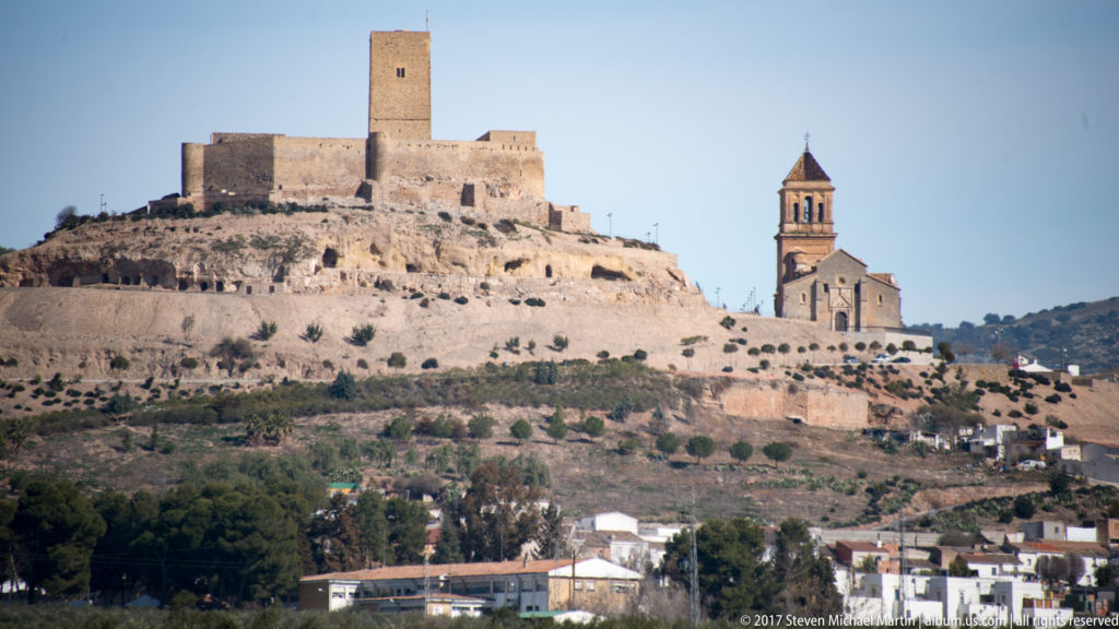 Rectangular castle keep and neighboring church bell tower on a rocky ridge above a town near Granada.