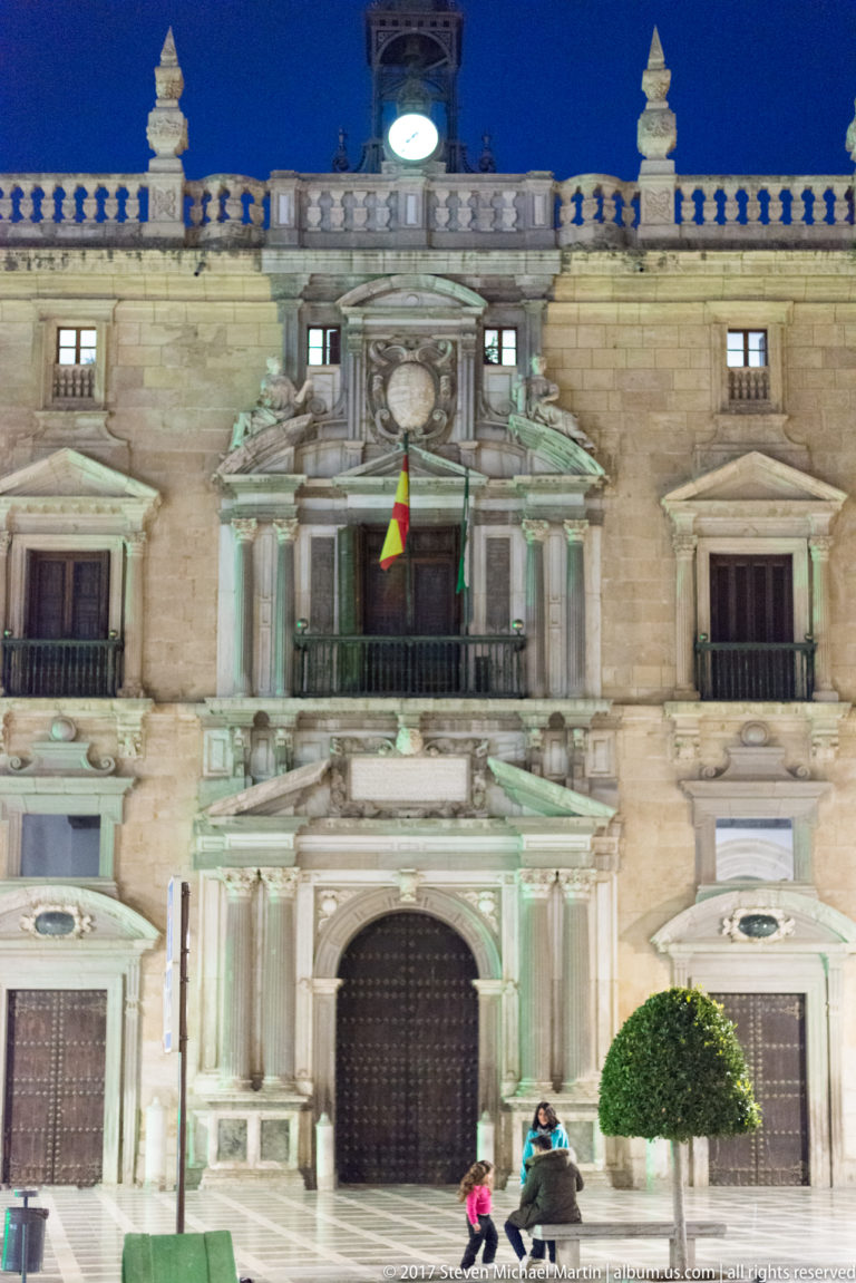 Central stone portal and balcony of an ornate Granada civic building at night, with Spanish flag and pedestrians.