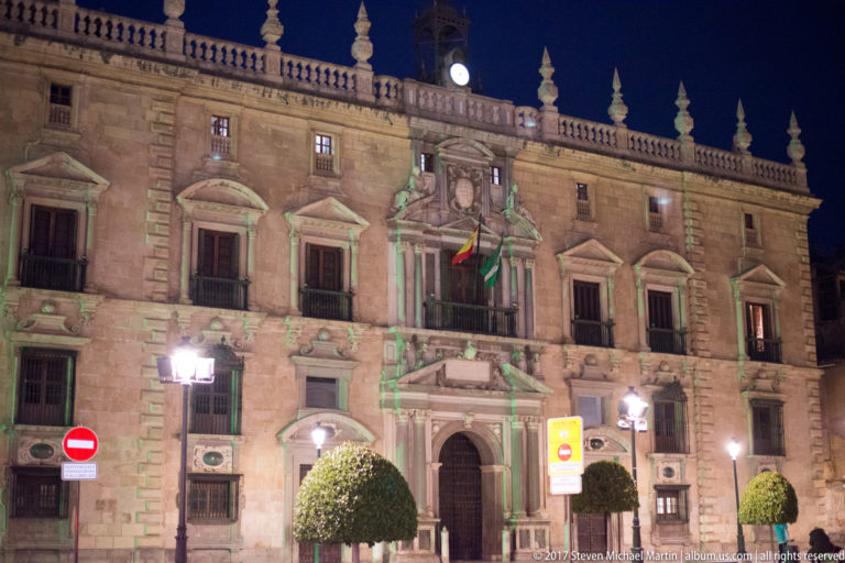 Illuminated Renaissance civic building with ornate portal, balconies, flags, clock, and clipped trees in Granada.