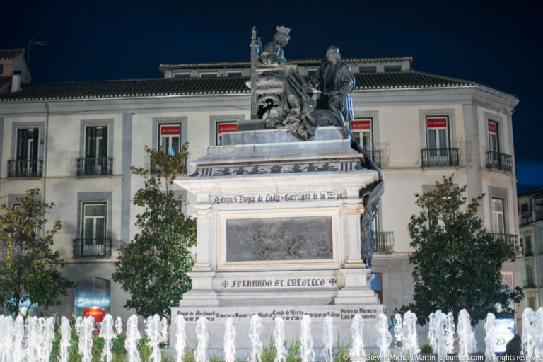 Isabella I and Christopher Columbus monument behind illuminated fountain jets and city façades at night.