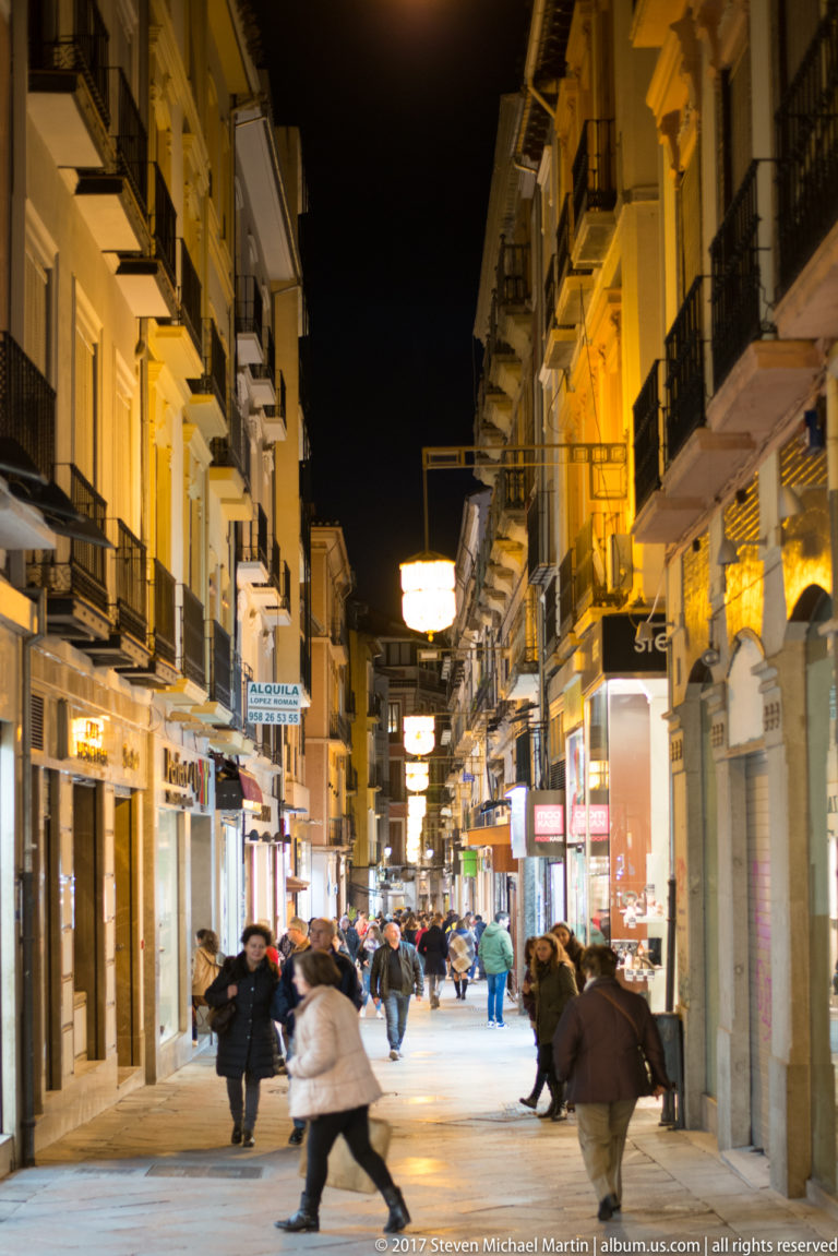 Pedestrians walking along a narrow, brightly lit Granada shopping street at night.