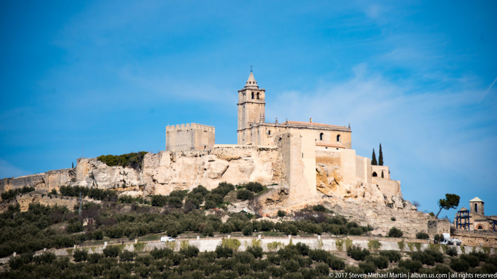 Stone hilltop fortress with square tower and adjoining church bell tower above olive-covered slopes near Granada.