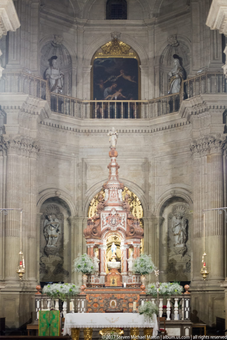 Polychrome altar centered beneath a high tribune, stone arches, reliefs, and painting inside Granada Cathedral.