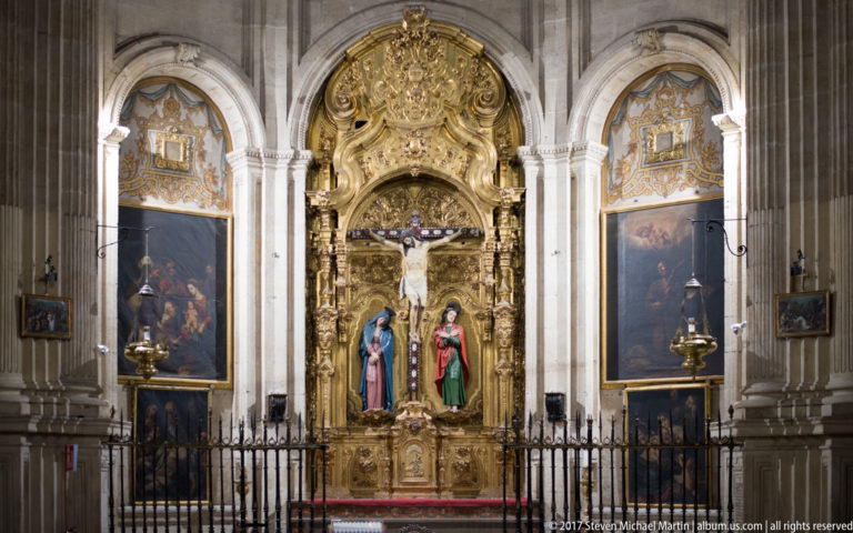 Gilded chapel retable with Crucifixion and two standing saints, flanked by paintings inside Granada Cathedral.