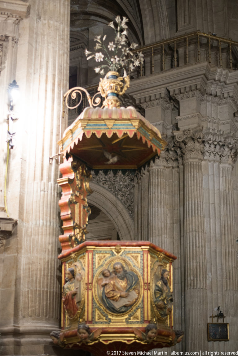 Colorful carved wooden pulpit with relief panels and canopy attached to a stone pier in Granada Cathedral.