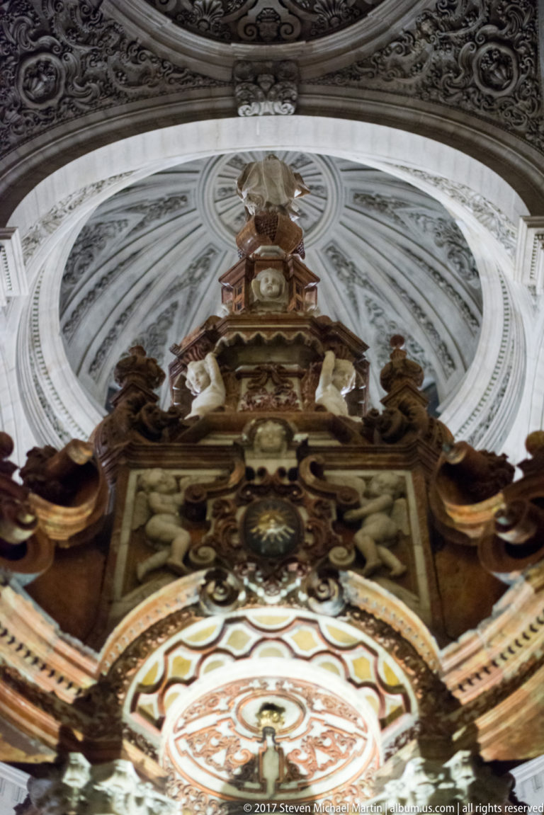 Upward view of carved altar crest with figures and scrollwork beneath the pale vault of Granada Cathedral.