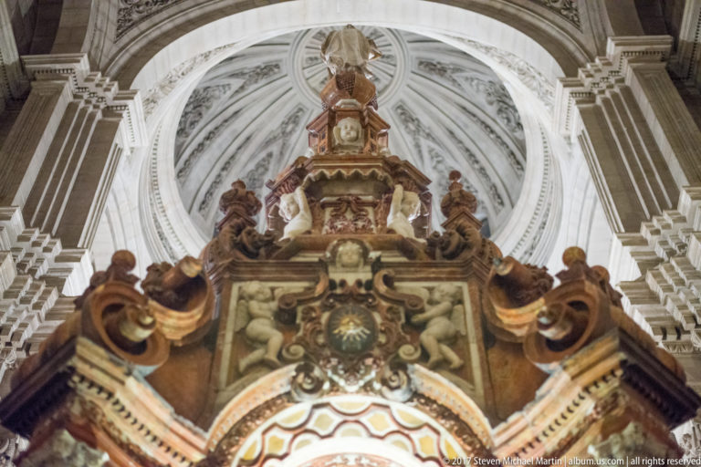 Ornate sculptural altar crown viewed from below between pale piers and radial vaulting in Granada Cathedral.