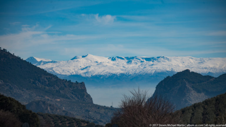 Snowy Sierra Nevada framed between dark rocky foothills under a blue winter sky near Granada.