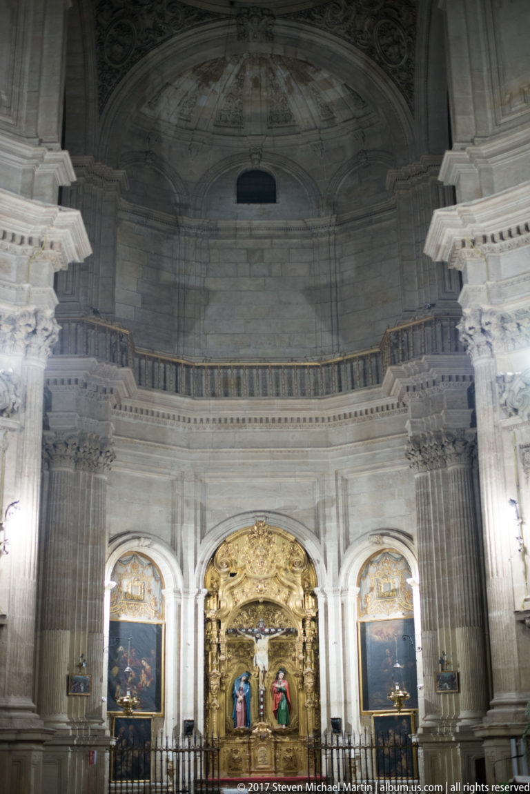 Distant gilded chapel centered beneath a towering curved stone wall and upper gallery in Granada Cathedral.