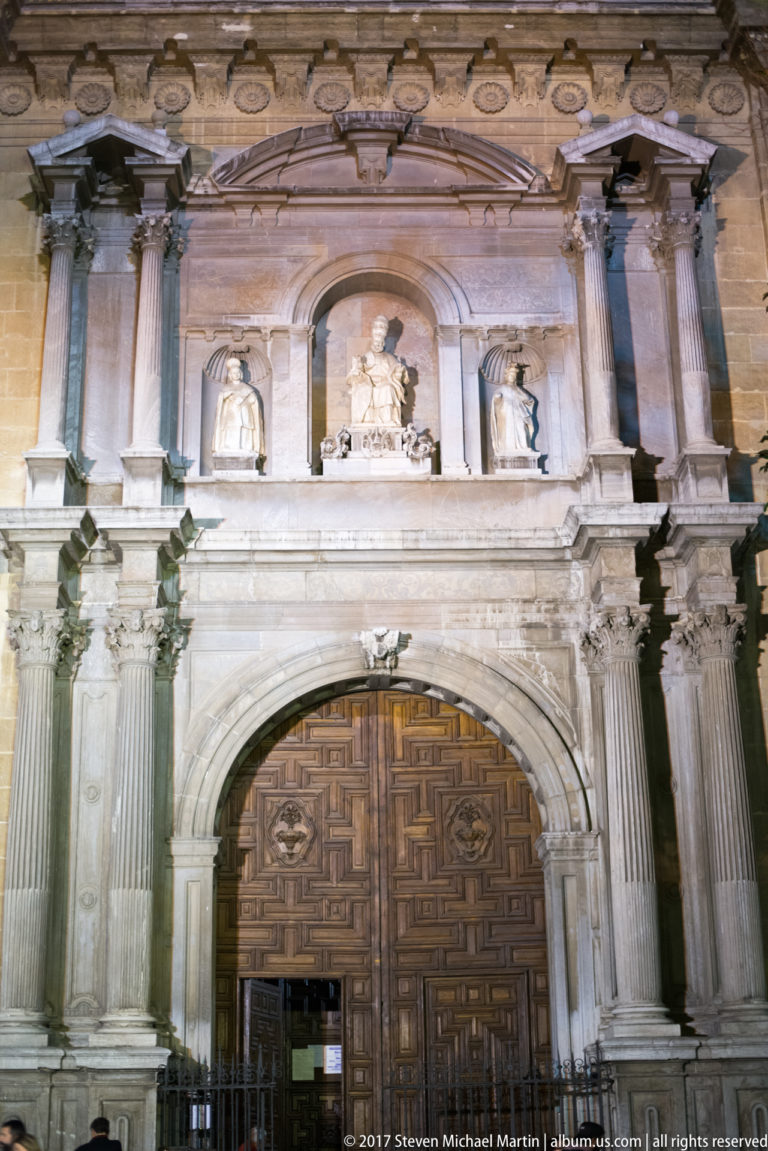 Illuminated monumental portal of Granada Cathedral with paired columns, carved niches, heraldry, and paneled wooden doors.