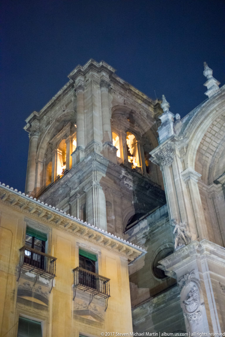 Illuminated stone tower of Granada Cathedral rising above neighboring façades under a dark blue night sky.