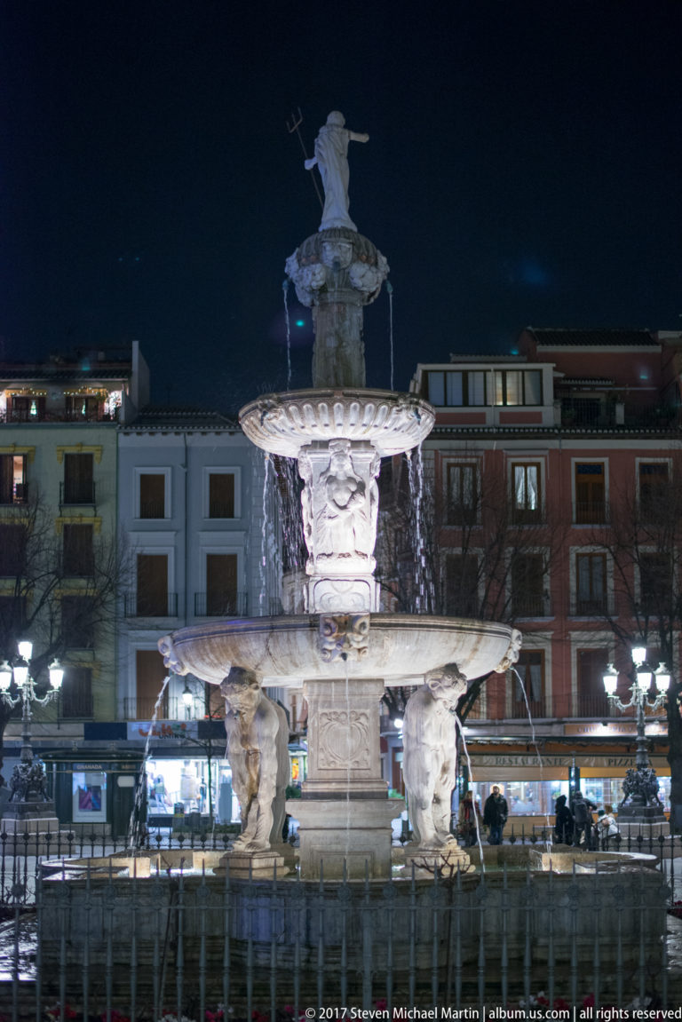 Illuminated multi-tiered Fuente de las Batallas with sculpted figures and flowing water in central Granada at night.