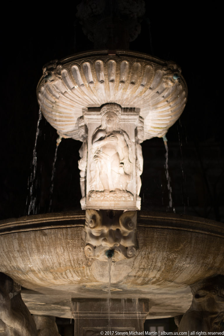 Illuminated upper basin and carved central support of Fuente de las Batallas with water streaming from the rim.