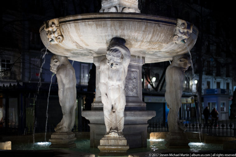 Three carved male figures supporting a broad stone basin on Fuente de las Batallas in Granada at night.
