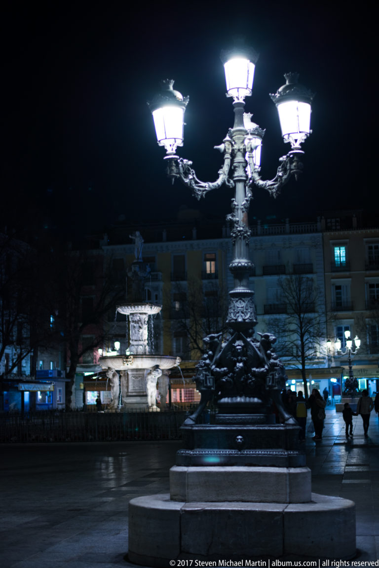 Ornate multi-lantern streetlamp glowing in a Granada plaza at night, with an illuminated fountain behind.