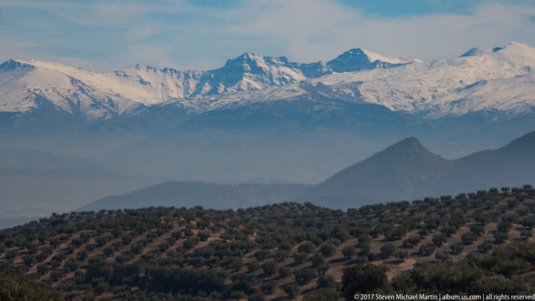 Snow-covered Sierra Nevada rising beyond hazy foothills and patterned olive groves near Granada.