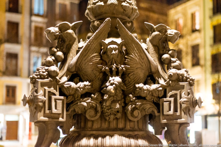 Close-up of a winged griffin-like figure, fruit, masks, and scrollwork on an ornate Granada streetlamp base.