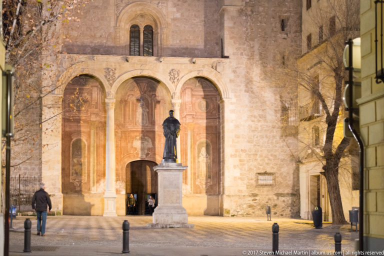 Bronze statue before a floodlit stone church with three tall arched portals in a Granada square at night.