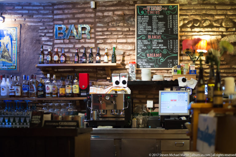 Compact Granada bar with bottles, hanging glasses, espresso machine, brick wall, and handwritten wine lists behind the counter.