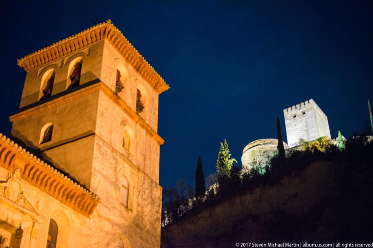 Illuminated church tower in foreground with Alhambra towers and walls rising on the hillside behind at night.