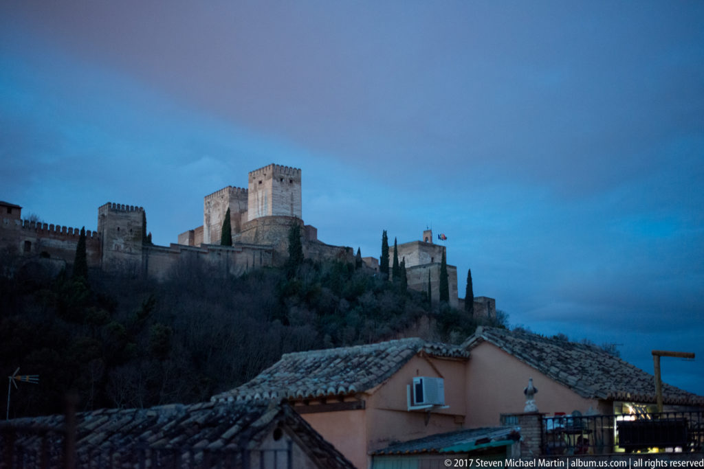 Alhambra walls and square towers extending across a wooded ridge above tiled Granada rooftops at dusk.