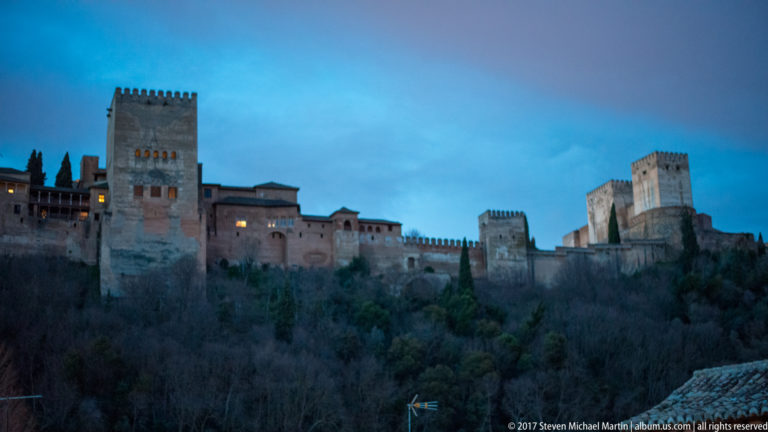 Alhambra towers, crenellated walls, and long ranges seen across a wooded slope during blue hour.