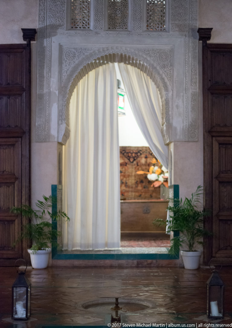 Ornately carved horseshoe arch with white curtains, dark wooden doors, and potted plants in a Granada interior.