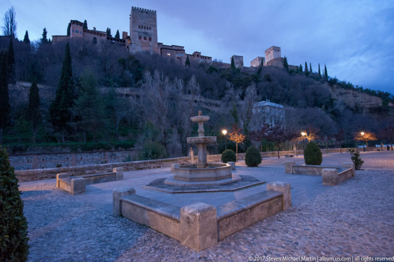 Two-tiered stone fountain in a paved Granada plaza with the Alhambra rising on the wooded hillside behind.