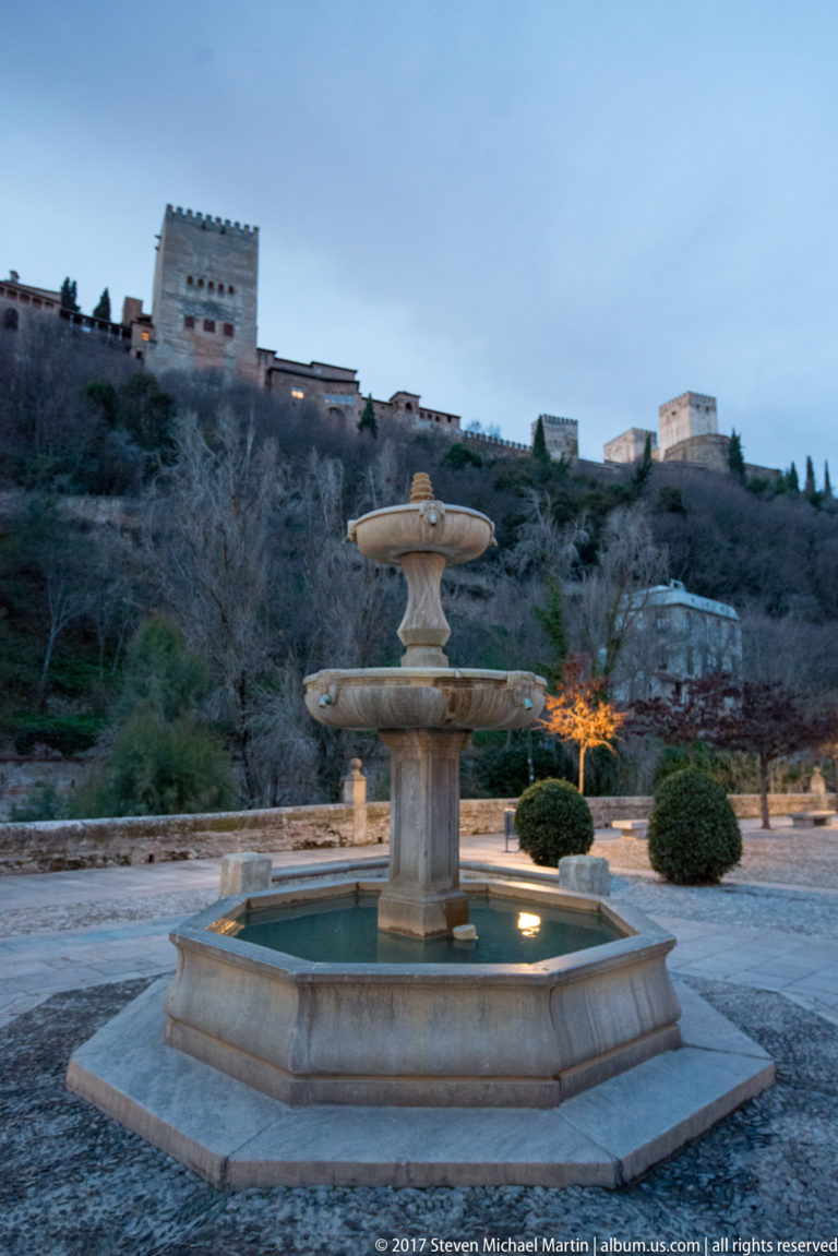 Close view of a two-tiered stone fountain with the Alhambra partly visible on the hillside behind.