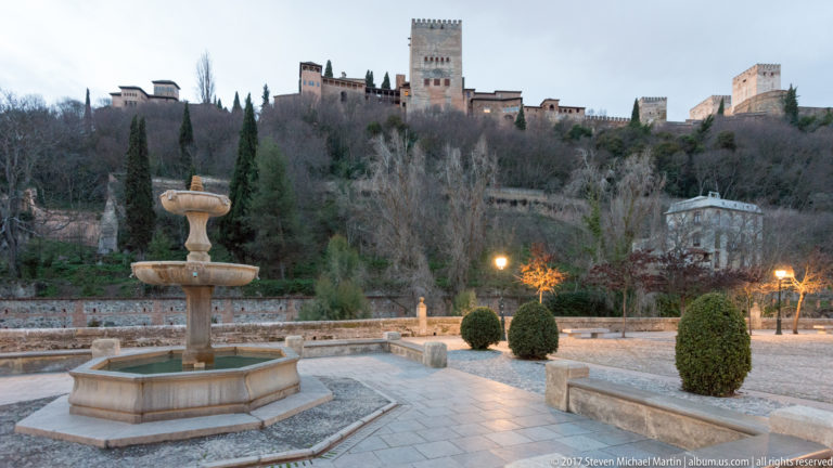 Stone fountain beside clipped shrubs and lamps, with the Alhambra stretching across the hillside above.