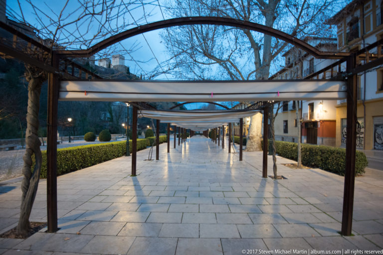 Long paved Granada walkway beneath a metal pergola, with clipped hedges, bare trees, and low buildings.