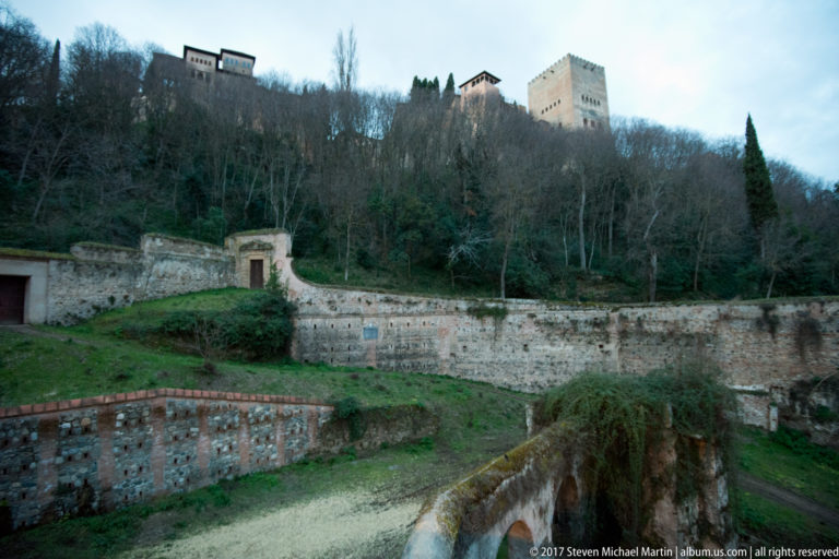 Weathered walls and narrow watercourse below two Alhambra towers on a wooded Granada hillside.