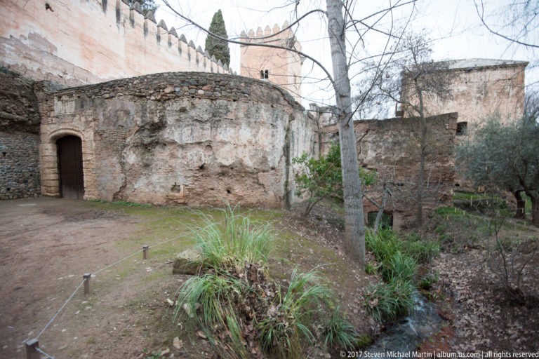 Weathered low masonry structure with arched doorway, exposed brick, and a small stream below the Alhambra hill.