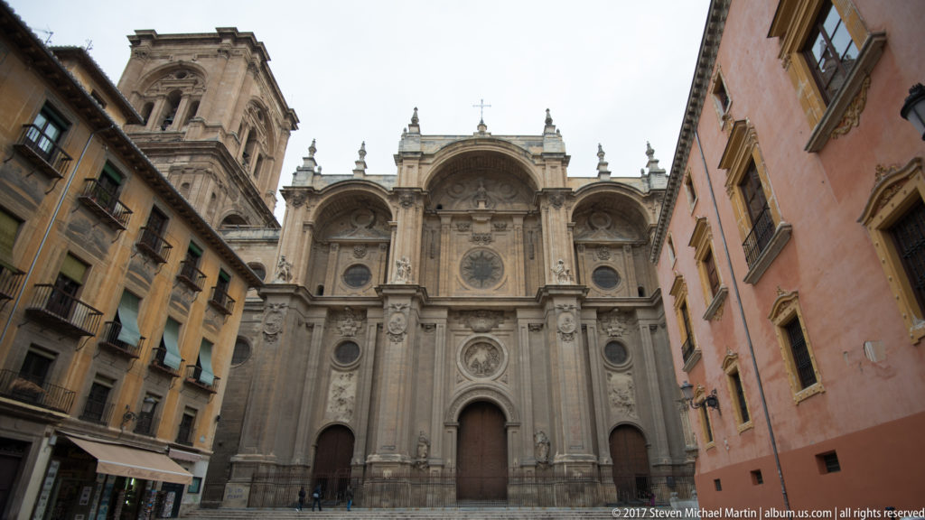 Granada Cathedral main façade framed by multistory buildings on both sides of a narrow urban plaza.