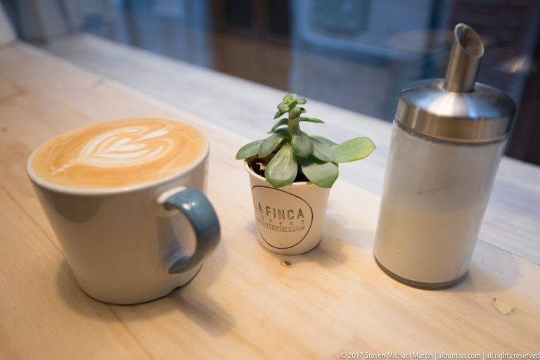Latte with leaf foam art beside a small succulent and metal sugar dispenser on a wooden café table.