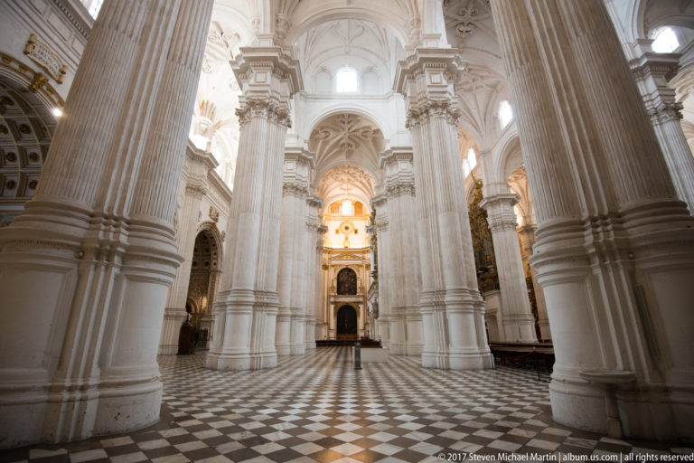 Wide interior of Granada Cathedral with massive white fluted piers, patterned floor, high vaults, and distant altar.