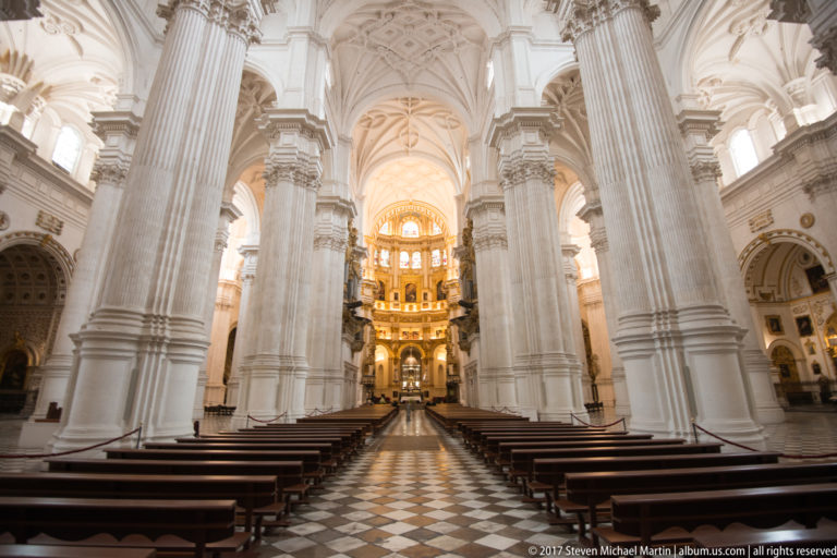 Central aisle of Granada Cathedral lined with wooden benches and massive white piers, facing the illuminated main chapel.