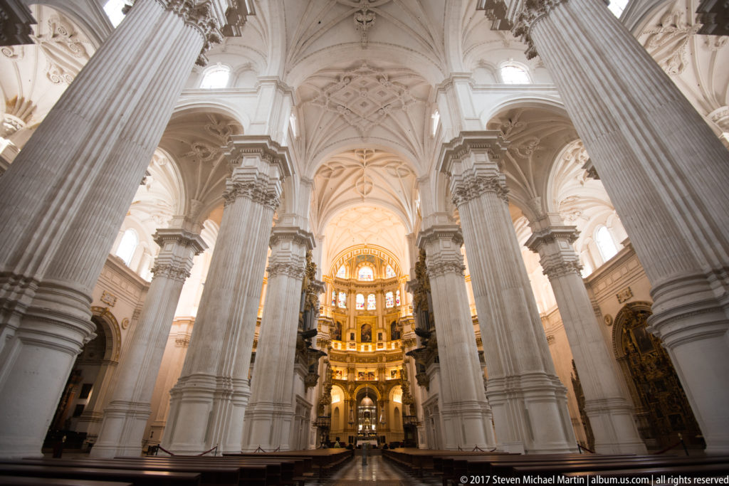 Granada Cathedral's illuminated Capilla Mayor framed by massive white piers, arches, and vaulted ceilings.