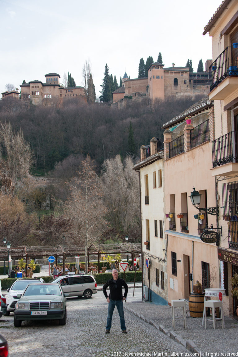 Cobbled Granada street with parked cars and pedestrian below the Alhambra complex on a wooded ridge.