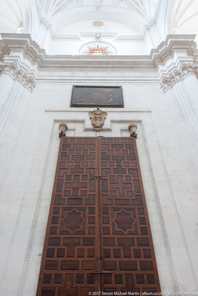 Large geometric-paneled double wooden door beneath a carved crest and plaque inside Granada Cathedral.