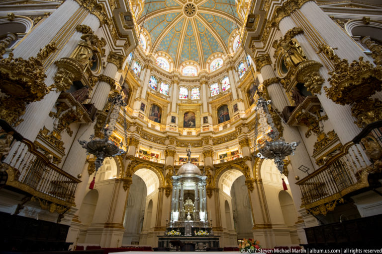Low-angle view of Granada Cathedral's Capilla Mayor, central sanctuary, stained glass, and blue ribbed dome.