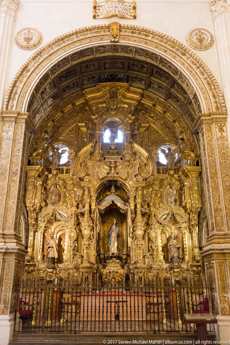 Highly gilded Baroque retable with sculpted saints and central devotional image behind an iron screen in Granada Cathedral.