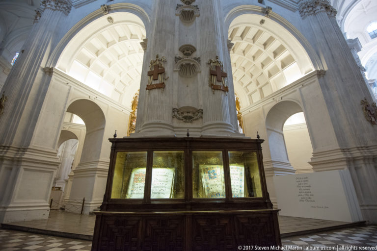 Wooden display case with illuminated historic pages standing beneath massive white arches inside Granada Cathedral.