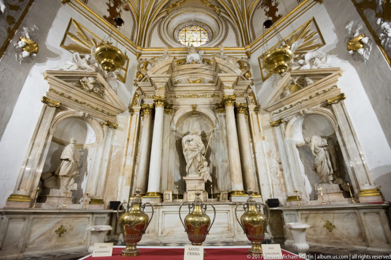 Marble cathedral altar with central saint, two flanking statues, dark columns, gilded capitals, and decorative vessels.